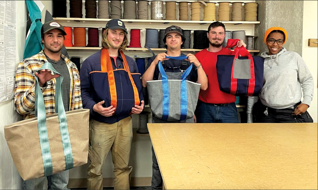 Five young adults pose with colorful tote bags in a workshop filled with spools of fabric. 