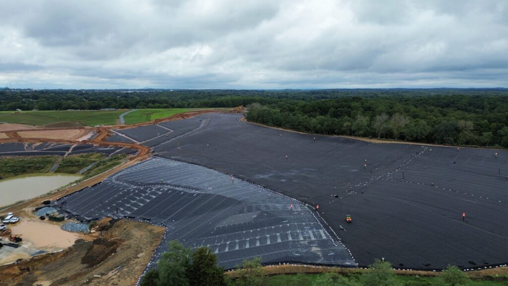 Aerial view of a landfill rehabilitation site with large black geosynthetic materials, workers, and green landscape in the background.