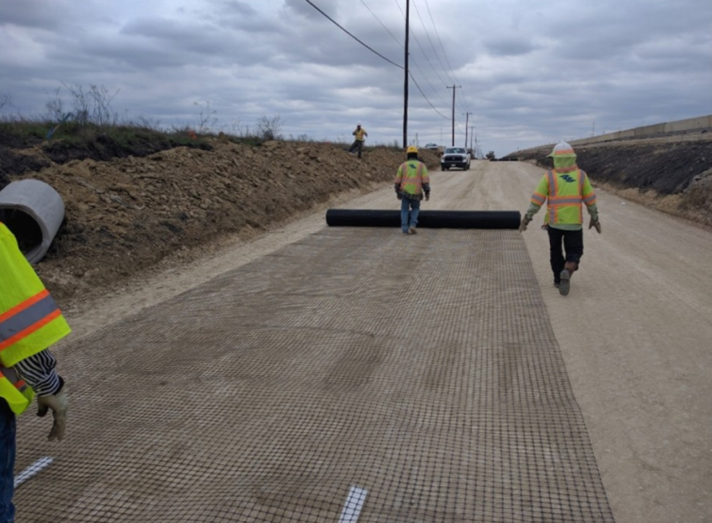 Construction workers in high-visibility gear lay a geosynthetic reinforcement mat on a dirt road, with power lines and cloudy skies above.