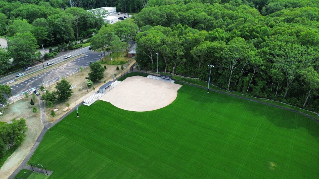 Aerial view of a recreational baseball field with lush green grass, a sandy infield, surrounded by trees and a parking area.
