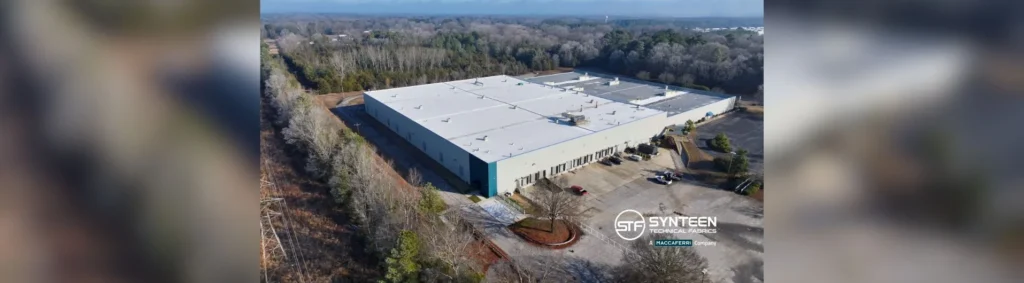 Aerial view of a large industrial building with a flat gray roof, blue accents, surrounded by trees and a parking area.