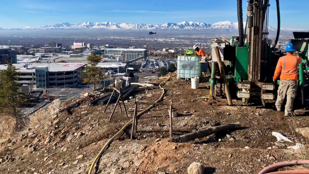 Construction workers in bright orange and blue helmets operate machinery on a hillside, overlooking a cityscape and snow-capped mountains.