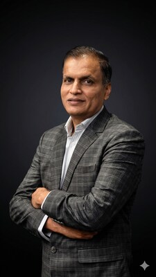 A man in a checkered gray suit with a white shirt stands confidently with arms crossed against a dark background.